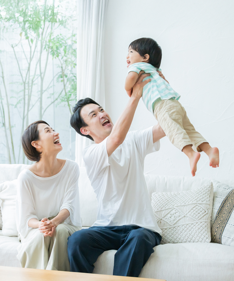 A couple sitting on a sofa, with the father lifting their child high, while the mother smiles at them, showcasing a joyful and warm family moment.