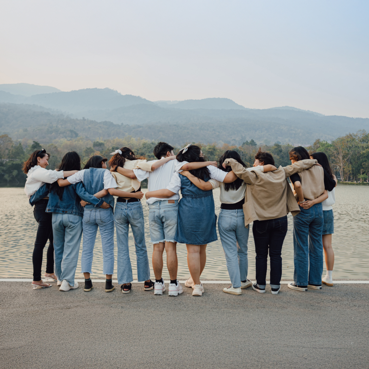 A group of young women and men stand by a lake, facing away from the camera, embracing each other's shoulders with mountains and the lake visible in the background.