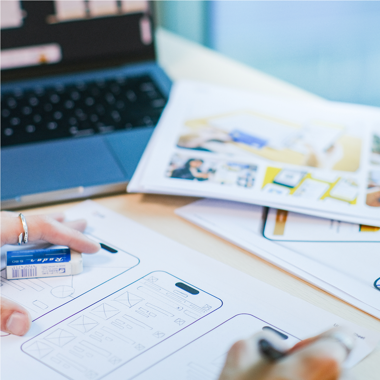 A close-up of hands working on wireframe designs for a mobile app. A laptop is visible in the background, along with printed materials and sketches on a table.