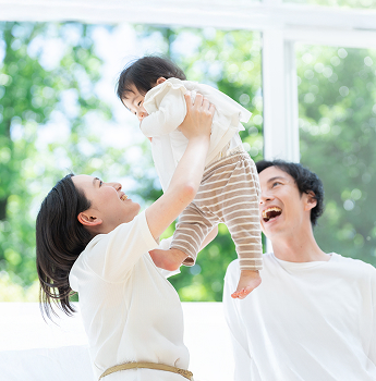 A couple in a bright indoor setting, with the mother lifting their baby high, while the father smiles at them, showcasing a warm family atmosphere.