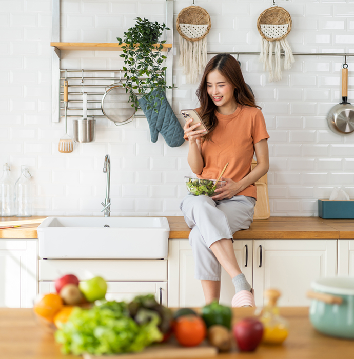 A woman sitting in a kitchen, holding a smartphone, with fresh vegetables and fruits nearby, showcasing a healthy lifestyle.