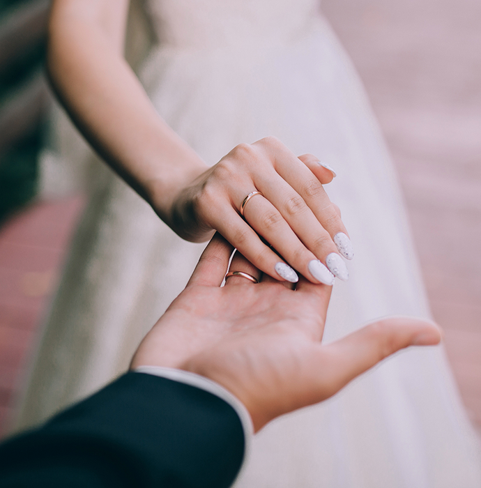 A close-up of a bride's hand holding the hand of a groom, showcasing a wedding band and elegant nails