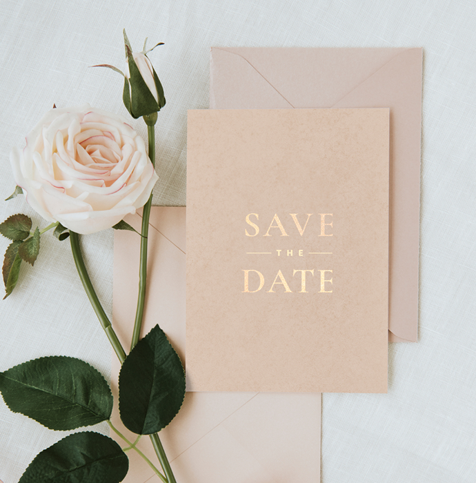 A flat lay of a “Save the Date” card with elegant gold lettering, accompanied by a soft pink rose and green leaves, resting on a light fabric surface.