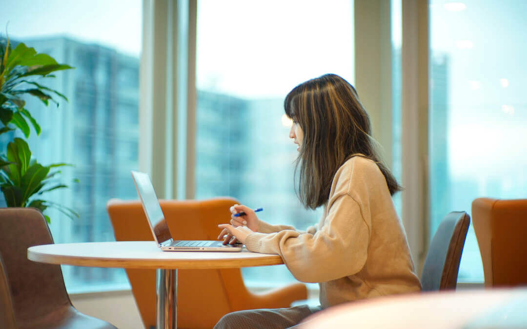 A woman is sitting at a round table facing a laptop, holding a pen in her hand and focusing on her work. The background features a cityscape outside the window.